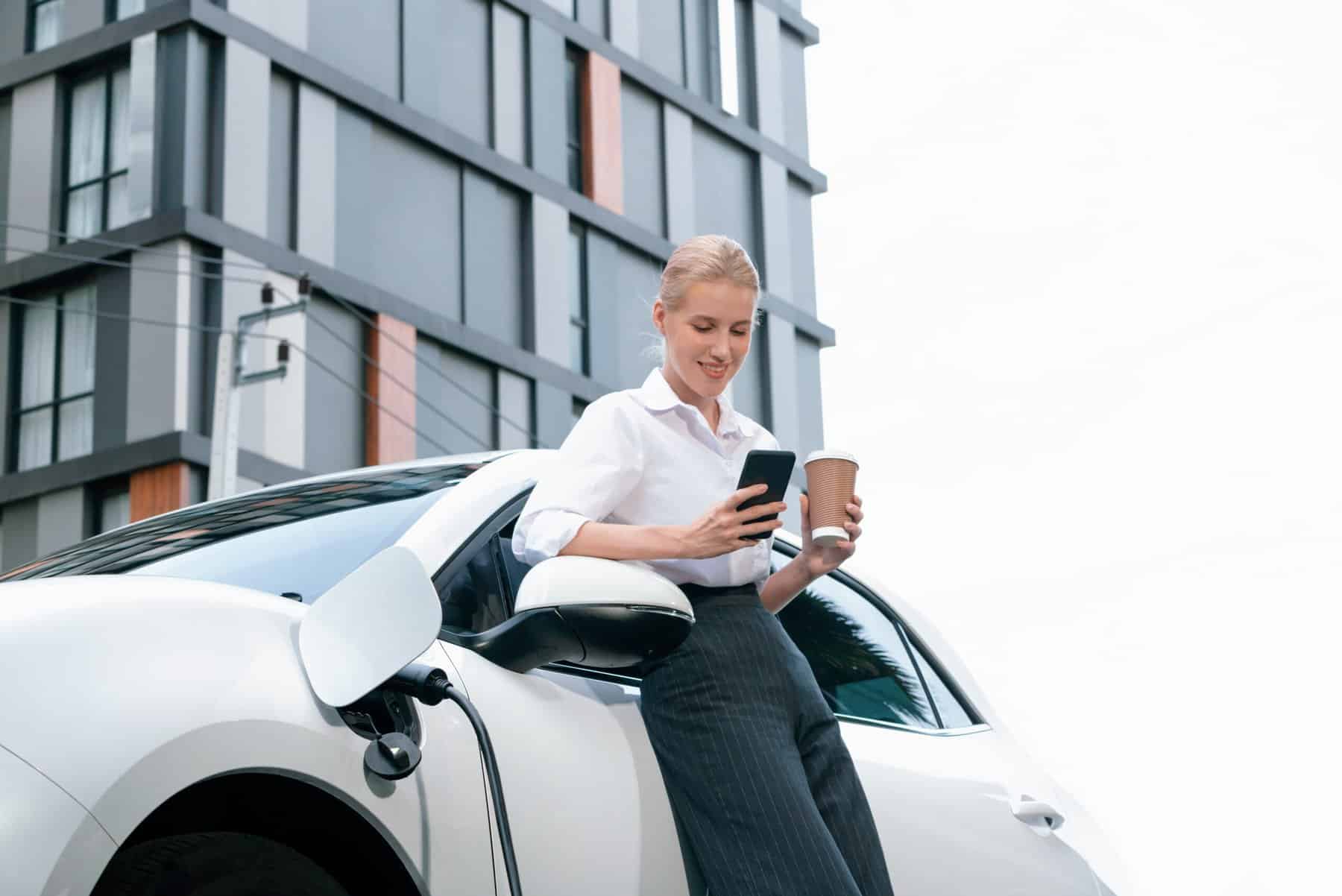 Lady is leaning against her white car which is plugged into an EV charger. She is holding her phone and a coffee and a tall office building is in the background.